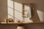 Beige bathroom wall with warm morning sunlight and soft shadows. A wooden wall rack holding a light beige towel draped naturally, matching the warm tones of the scene. Same woven accessories and basket below.