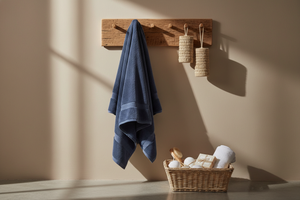 Beige bathroom wall with warm morning sunlight and soft shadows. A wooden wall rack holding a navy blue towel draped naturally, with two beige woven bath accessories beside it. Spa-style setup with a basket of bath items at the bottom. Same composition, same lighting, ultra-realistic.